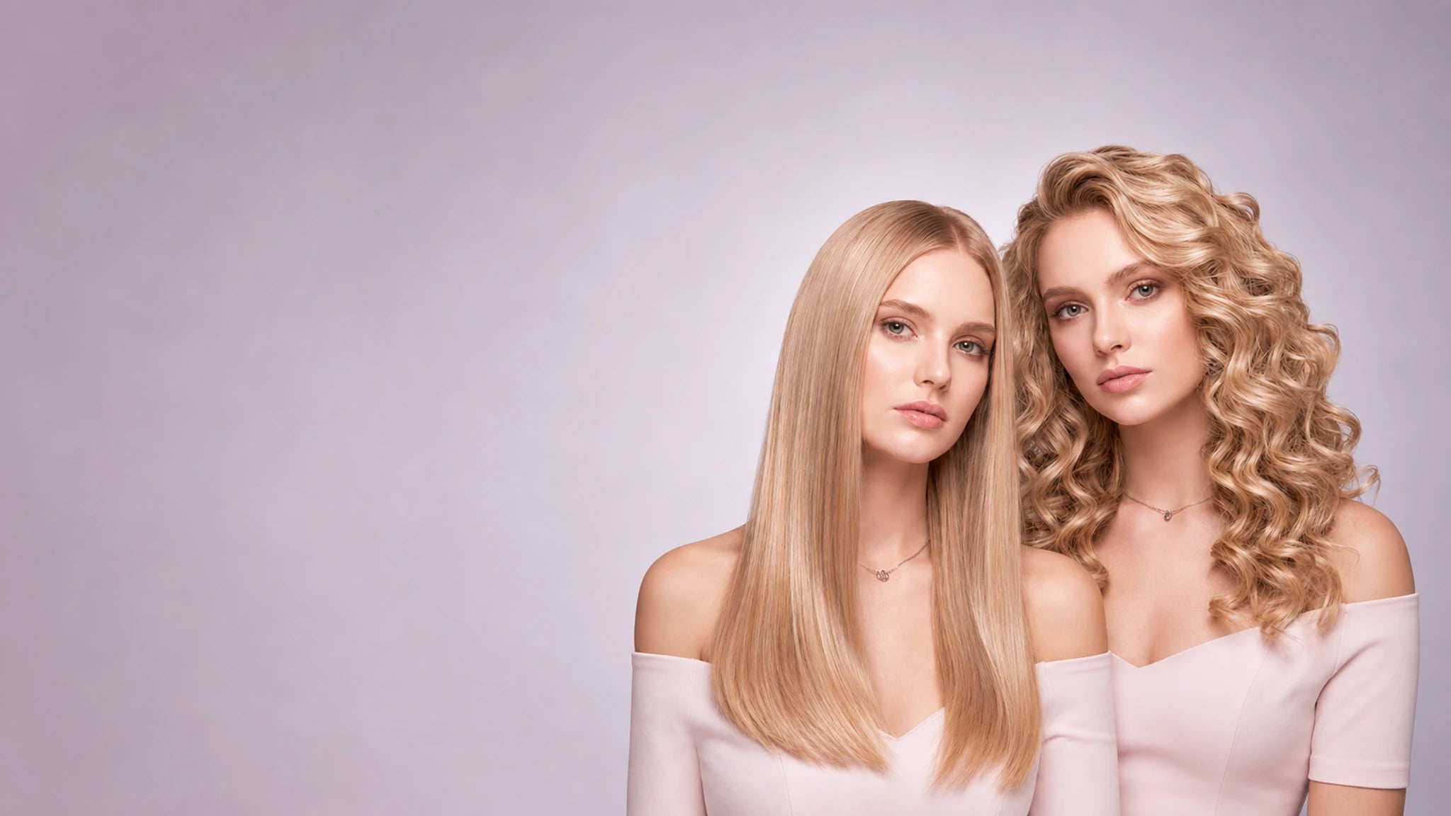 Two women posing side by side showcasing different hair types: one with straight, shoulder-length blonde hair and the other with voluminous, curly blonde hair. Both are wearing off-the-shoulder light pink tops against a solid lavender-gray studio background.